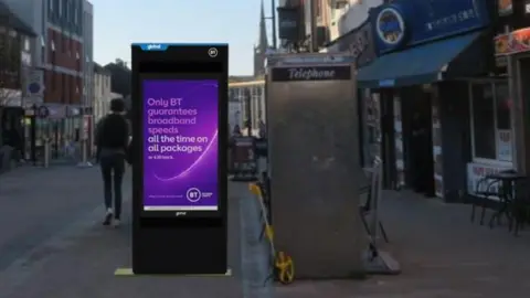 BT An artist's impression of a large black digital unit with a purple screen on it. It is next to a phone box on a pedestrianised street. 