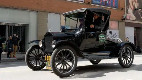 NurPhoto vía Getty Images Un automóvil Ford Modelo T circula por una carretera. Una pareja parada en la acera mira pasar el coche. Uno de ellos le toma una foto con su teléfono. 