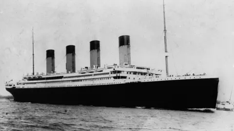 A black and white photo of the Titanic on the open water. It has four large funnels and its hull is dark black