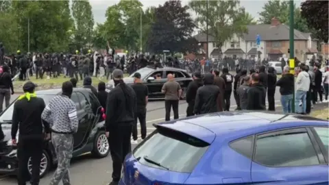 Crowds gather on a street near a McDonald's close to the Heartlands Hospital, with three cars in the shot
