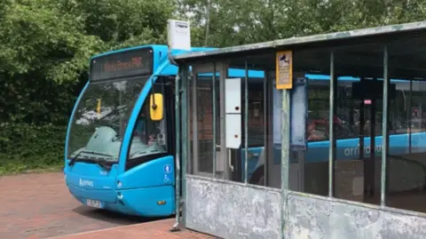 BBC A blue bus parked behind a dirty metal bus shelter on a red-coloured road