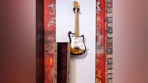 The Beatles Story/PA Media George Harrison's Futurama electric guitar hanging up on a white board on a wall along with an old guitar case and the red wall littered with graffiti 