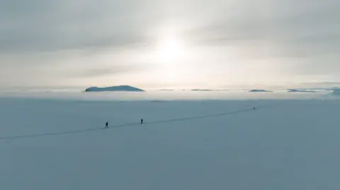 Beyond the Ultimate A wide aerial shot of the Arctic environment. It is a vast open space of land completely covered in snow and ice, with bits of cloud above it. Through the middle is a path trodden by people, with three participants trekking along it. There are large mountains peeking above the clouds