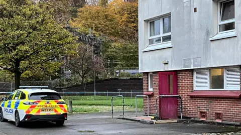 Exterior of tower block in the early morning with one police car parked outside and police tape cordoning off an area in the front corner of the building