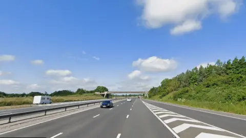 The photo shows a section of motorway as seen from a car. The beginning of a slip lane with black and white arrow markings can be seen to the right and there is a slim concrete bridge passing over the motorway in the background. The sky is blue and dotted with clouds.