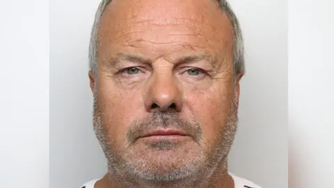 Jack McDaid, a man in his 60s with short grey hair wearing a white and black striped T shirt, standing against a grey background.