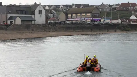 A modern orange lifeboat with three people on it all in protective gear travels out and along the beach. In front of them is the beach and the beach amusements.