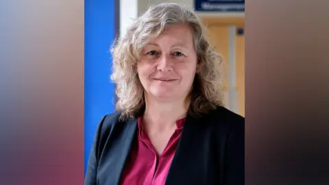The QEH Jo Segasby has shoulder-length greying brown hair. She is wearing a black jacket with a dark pink shirt underneath