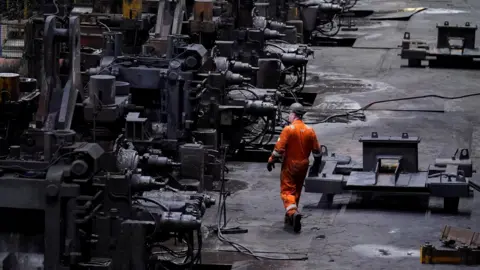 A wide view inside an industrial steelworks, showing long rows of large, dark metal machinery and workstations. A worker in bright orange protective clothing walks along the factory floor between the machines, with cables, metal parts, and heavy equipment scattered across the ground under dim industrial lighting.
