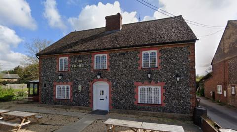 The outside of The Chequers, a pub with a pebbledash exterior, five windows and a white front door. There is a chimney in the middle of the roof and it looks like it's in a rural setting. There are picnic benches at the front
