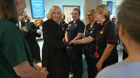 Queen Camilla is inside a hospital ward surrounded by female hospital staff. She is in the middle of a conversation with them.