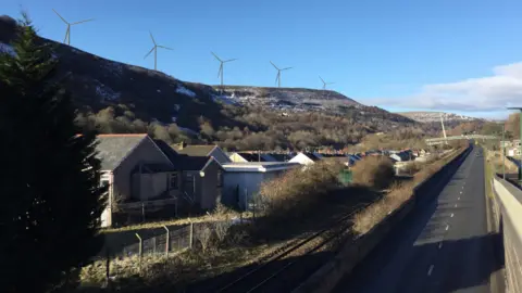 Cenin A mock up of a road and railway line next to a row of terraced houses in front of some steep hills in the background. On the stills there are five impressions of wind turbines 