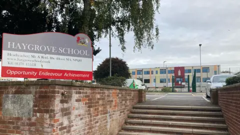 Haygrove School sign with a red brick wall. There's steps up towards a car park and then a building in the background.