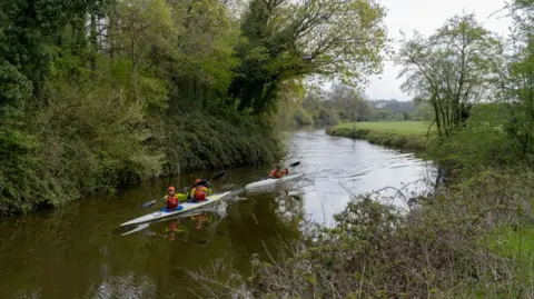 A stock image of people kayaking down the river, there are three people in two boats. They are wearing Hi-Viz jackets. The weather is dull and there is lots of greenery around. 