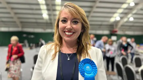 Sarah Bool is standing in the middle of the picture smiling. She has a white blazer on, with a navy top underneath. She has ginger hair, and is wearing a blue rosette with her name on. 