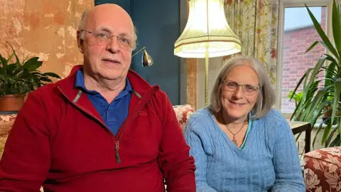 A man and a woman sat on a sofa. The man is bald and wears glasses and a red jumper. The woman wears glasses and a blue knitted jumper.
