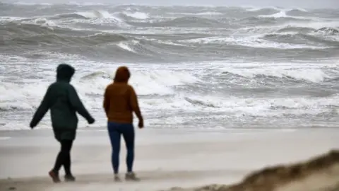 EPA Two women walk on a beach as waves crash on to the sand. 