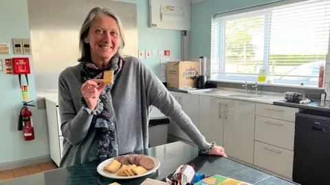 Warm hub organiser Paula Morton wears a grey jumper and a floral scarf. She is standing in the village hall kitchen holding a shortbread biscuit from a plate in front of her, which is on a table. She has shoulder length, grey hair and is smiling at the camera. To her left is a sink, cupboards and windows. 