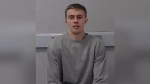 A young man with short brown hair, pictured in a grey sweatshirt against a grey wall