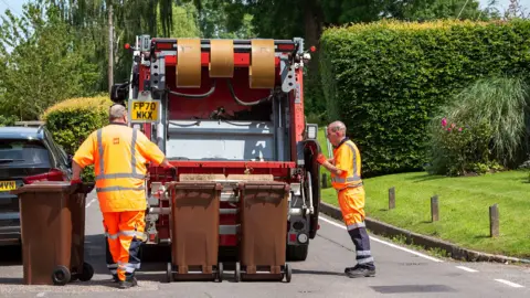 Getty Images A rubbish colection truck in Hampshire is seen from behind with two refuse collectors wearing orange overalls . It is a bright and suuny day 