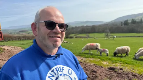 A farmer standing in a blue and white branded polo neck top and jumper with Cantref Farm written on it. He is looking towards the camera on a sunny day as some of his sheep graze in a field behind him.
