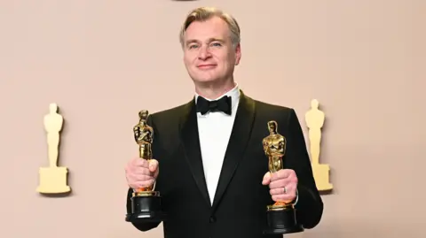Getty Images Christopher Nolan smiling successful  a achromatic  bow necktie  and meal  jacket, holding an Oscar statuette successful  each   hand, successful  beforehand   of an Oscars backdrop