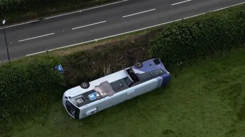 A purple and white bus is seen on is side in a field