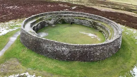 A stone ring fort sitting on a grassy mound, surrounded by brown gorseland 