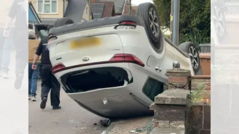 A white Kia on its roof partially on the pavement and partially in the road after crashing. Debris and smashed glass can be seen on the floor.