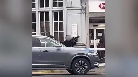 @ddotz6/TikTok An armed police officer crouching down behind a silver car outside a branch of HSBC bank, aiming a gun upwards