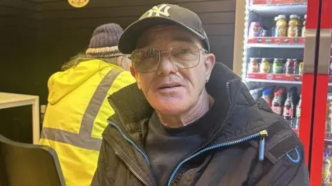 Ivan Millward has a black New York Mets baseball cap, black T-shirt, black anorak with light blue facings. He has short grey hair and is wearing glasses.