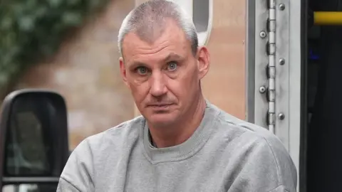 Donald Excell staring into the camera, he is in a grey prison issue jumper.