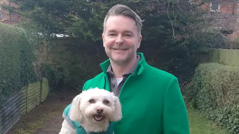 Chris is standing in a garden wearing a green coat, green jacket and a white top. He has black and grey hair and facial hair. He is holding a white fluffy dog with a green coat on it. Behind him are green hedges, a tree and grass.