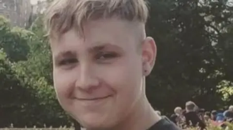 Police Scotland A young man with blond hair, wearing a dark grey t-shirt, smiles at the camera in a close-up shot with trees and greenery in the background. 