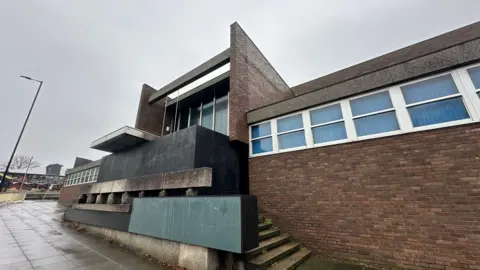 A general view of the front of the former crown court in Ipswich. It is a red brick building and its front entrance has been boarded up with large black boards. The windows on the building have blinds pulled across them. 