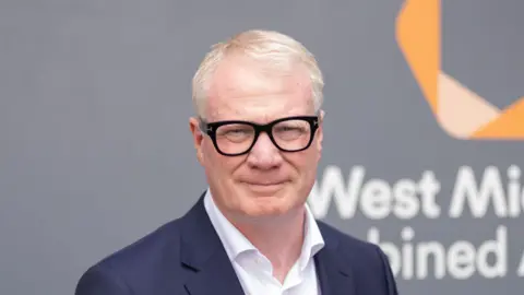 West Midlands Combined Authority Mayor Richard Parker wearing black rimmed glasses, a white shirt and dark blue blazer is looking at the camera with a slight smile. He is standing in front of a grey wall with the words West Midlands Combined visible over his right shoulder and a yellow twisted ribbon motif above the words