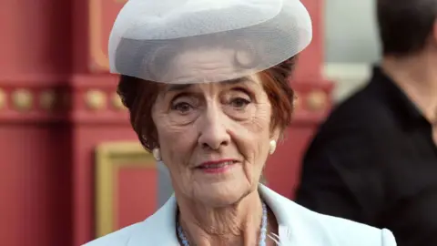 June Brown as Dot Cotton, a woman with short brown hair in a pale blue suit and hat, with blue beads, holding a small camera on the set of BBC soap Eastenders 