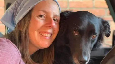 Family photo Jo is wearing a pink top and blue patterned headscarf and she has shoulder-length hair. She is smiling and her face is near a pet dog which is black with grey whiskers. 