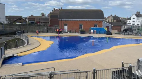 A paddling pool filled with water. The floor is electric blue with a yellow border. In the background are buildings and construction workers in hi-vis jackets. 