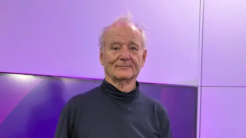 Bill Murray pictured in the BBC WM studio wearing a navy blue turtleneck jumper. He has thin, white hair. He is stood in front of a TV screen which has purple BBC WM branding on it. 