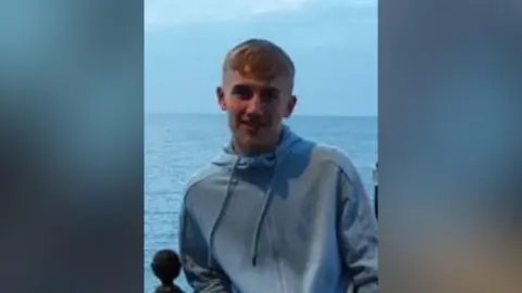 A young man in a pale blue hooded top standing on a sea front