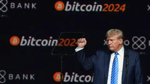 Donald Trump in a navy blue suit and royal blue striped tie holding his fist up during a Bitcoin-sponsored event at which he spoke. 
