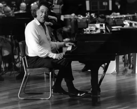 Getty Images A black and white image of Duke Ellington sitting at the piano in the cathedral. He is wearing a light shirt, dark trousers and shoes. 