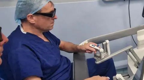 Royal Cornwall Hospitals NHS Trust A medic wearing blue surgical scrubs and a head covering is sitting at a screen which shows an operation. He is holding two controls to direct the operation.