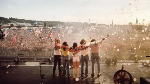 Supplied Five members of Wolf Alice stand on the stage after performing at Glastonbury. The photograph is taken from behind them. In the distance is a crowd of thousands of people 