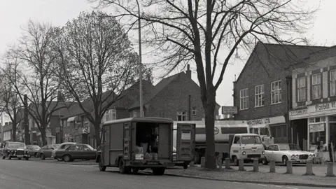 A black and white image of a van with open back doors, a tanker lorry and a row of shops near a road.