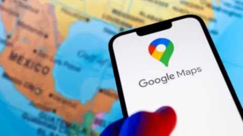 A hand holds a phone that displays the Google Maps logo in front of a map that shows Mexico and the US