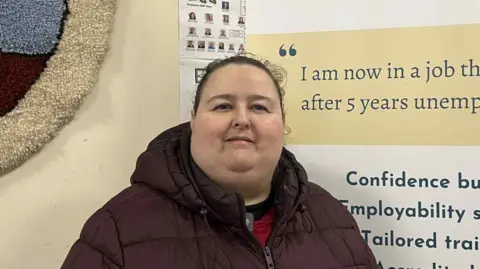 Patricia McKeag has dark hair tied up. She is wearing a burgundy quilted coat. Behind her is a sign with writing on it. The writing says "I am now in a job that I enjoy after 5 years unemployment." "Confidence building employment support tailored training Accredited training Goal setting"