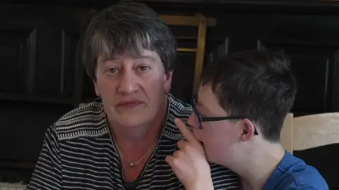 A woman with short brown hair wearing a black and white striped T shirt. Next to her sits a teenage boy with Down's syndrome wearing a blue T shirt and glasses.