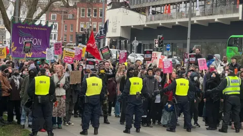 A large group of protesters in Bristol city centre, holding flags and placards. A row of police officers in high-vis vests stand in front of them.